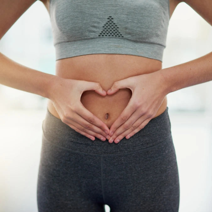 A person wearing a gray sports bra and gray leggings forms a heart shape with their hands over their stomach. The background is softly blurred.